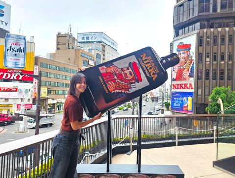 Woman poses with large Nikka Whisky bottle display in a city street.