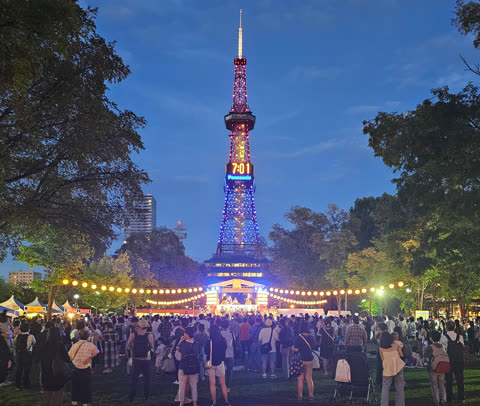 Nighttime crowd watching a performance in a park, illuminated tower in the background.