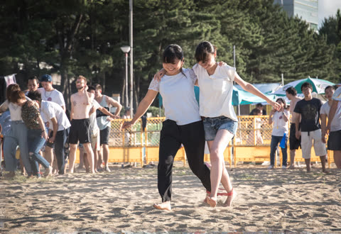 Two smiling women with their ankles tied together run in a three-legged race on a sandy beach surrounded by spectators.
