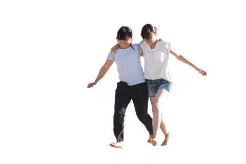 Two smiling women walk arm-in-arm with their bare feet lifted off the ground against a white background.