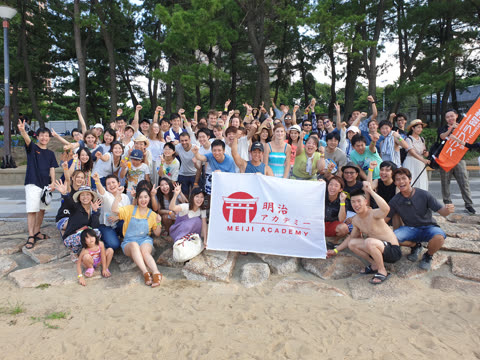A large group of people poses together for a photo on a sandy beach, holding a white Meiji Academy banner.