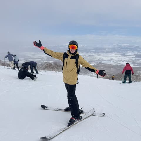 A smiling person in full ski gear stands with arms outstretched on a snowy mountain slope overlooking a valley.