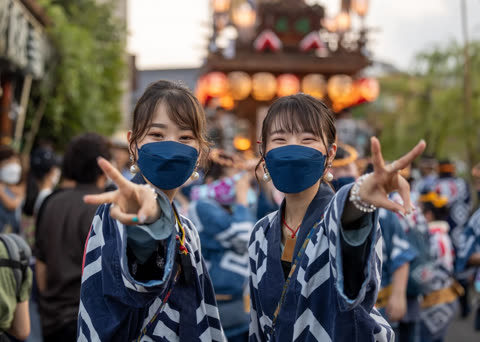 Two women in matching happi coats and face masks make peace signs at an outdoor festival.