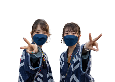Two women in patterned jackets and blue face masks make peace signs towards the camera against a white background.