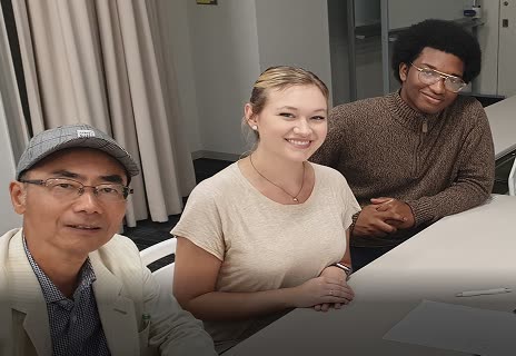 Three diverse people, an older man in a cap, a young woman, and a young man with glasses, smile while seated at a table indoors.