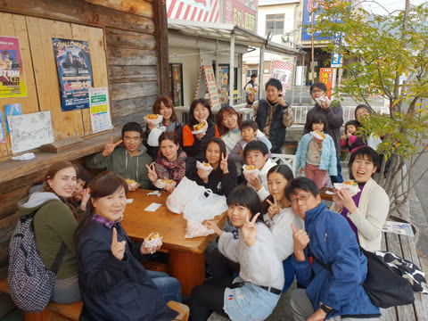 A large group of people smile and pose with food while gathered around an outdoor wooden table on a street in Japan.