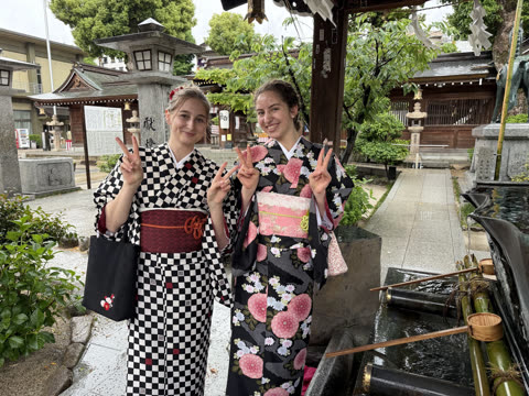 Two women in kimonos make peace signs while standing in the courtyard of a Japanese shrine.