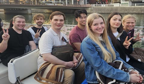 A group of seven people smiles and makes peace signs while sitting on a boat on a river in a city.