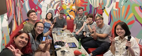 A group of people smiles and makes peace signs while sitting in a booth at a colorful karaoke room.