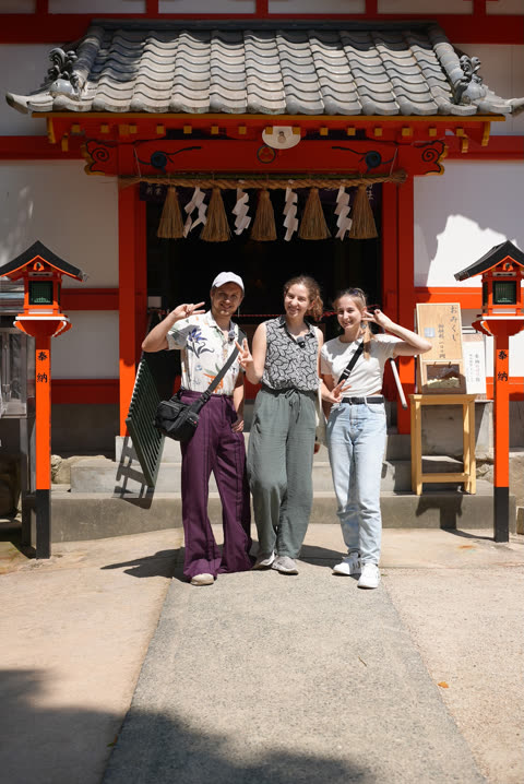 Three people making peace signs smile for a photo while standing in front of a Japanese shrine.