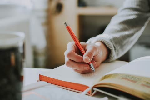 Person's hand writing with a red pen in a notebook.