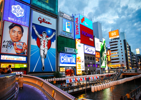 Tall buildings covered in large, illuminated advertisements with Japanese text tower over a river and a pedestrian bridge at dusk.