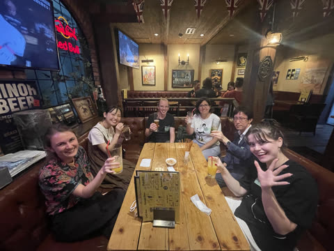 Six people sit together at a wooden table in a pub, smiling at the camera and holding their drinks.