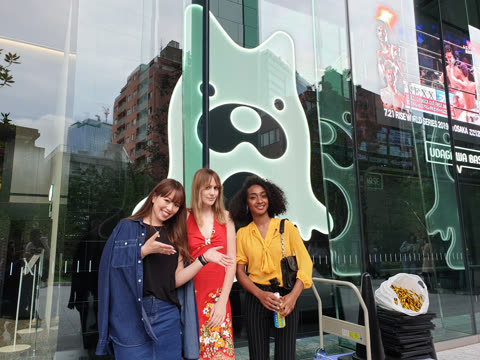 Three women pose together for a photo on a city sidewalk in front of a glass building with a large, glowing figure.