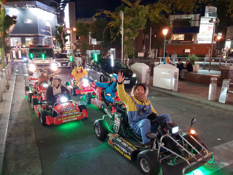 People wearing costumes drive go-karts in a line down a city street at night, as the driver in front waves.