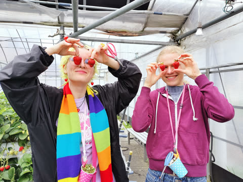 Two women hold strawberries over their eyes in a greenhouse.