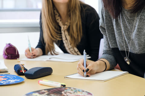 Two people write with pens in notebooks while sitting side-by-side at a wooden desk.