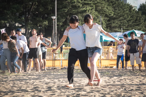 Two women walk barefoot on a sandy beach, their ankles tied together.