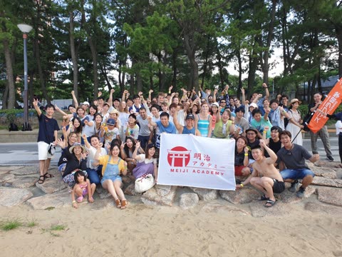 Large group posing happily by a beach, holding a Meiji Academy banner.