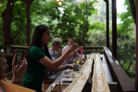 A woman lifts pink noodles from a flowing bamboo water chute with chopsticks while sitting on an outdoor porch.