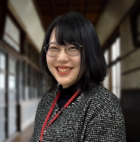 A woman with short black hair and glasses smiles while wearing a houndstooth coat in a long hallway.