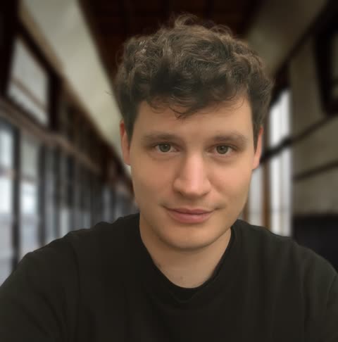 A man with curly brown hair wearing a black shirt looks at the camera in a long, out-of-focus indoor hallway.
