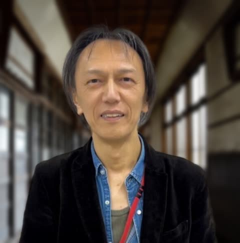 An East Asian man with dark, shoulder-length hair looks toward the camera while standing in a long, blurred hallway.