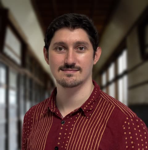 A man with dark hair and a goatee wearing a red patterned shirt looks at the camera inside a blurred-out hallway.