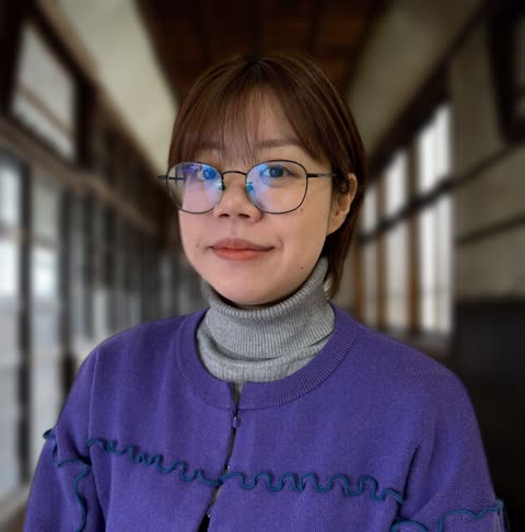A woman with short hair and glasses wearing a purple cardigan and grey turtleneck looks at the camera in a long room.