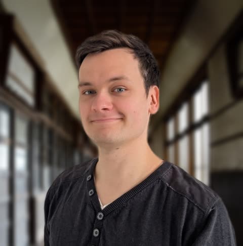 A young man with blue eyes and dark hair smiles slightly while standing in a long, windowed hallway.