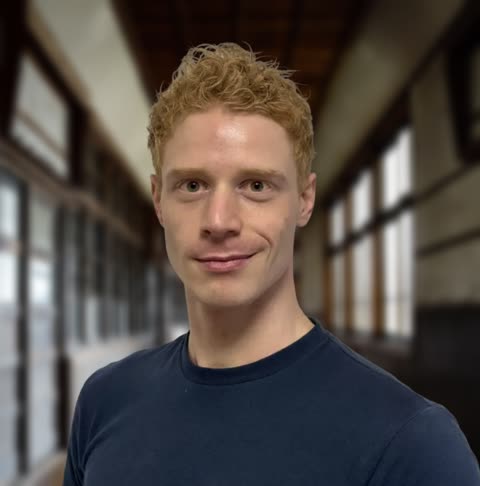 A man with curly blond hair in a blue t-shirt smiles slightly while standing inside a long, blurred hallway.