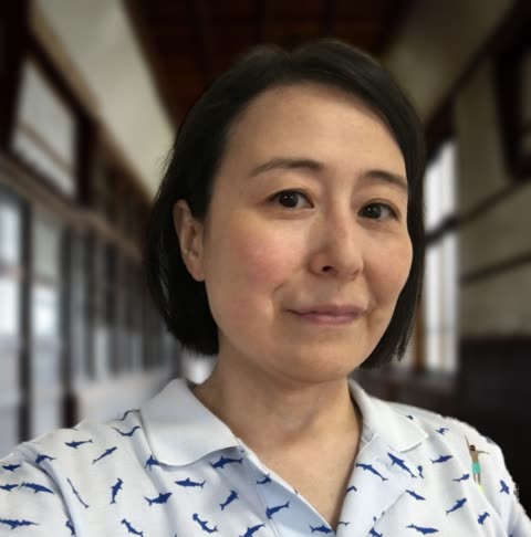 A person wearing a shark-print polo shirt smiles while standing in a blurred, window-lined hallway.