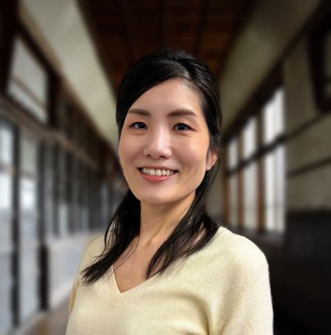 An East Asian woman with black hair in a cream sweater smiles at the camera while standing in a long hallway with windows.