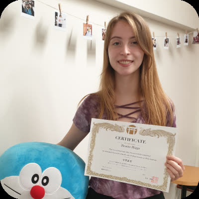 A smiling woman holds a certificate while standing beside a large blue plush toy in a room with photos on a string.