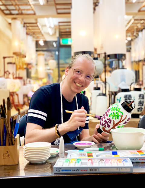 A person with glasses sits at a table and paints a colorful tree onto a paper lantern in a brightly lit workshop.